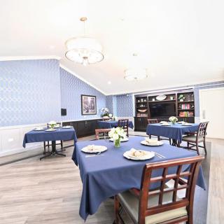 A bright dining room with blue tablecloths and neatly arranged tables and chairs, featuring elegant chandeliers and bookshelves.