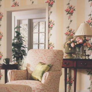 A cozy living room corner featuring a floral patterned armchair, matching wallpaper, and a decorative table with a lamp and flowers.