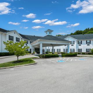 A large building with a covered entrance, surrounded by a parking lot, trees, and a clear blue sky with clouds.