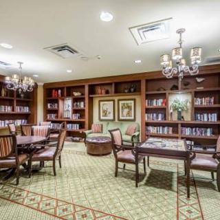 A cozy library room with bookshelves, tables, and chairs illuminated by elegant chandeliers.