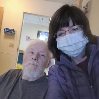 A selfie taken in a hospital setting, featuring an older adult and a caregiver, both wearing face masks.