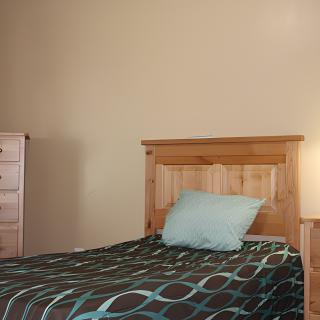 A simple bedroom with a single bed featuring a wooden headboard, accompanied by a dresser and a lamp.