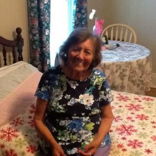 An elderly woman sitting on a bed, smiling in a cozy room.
