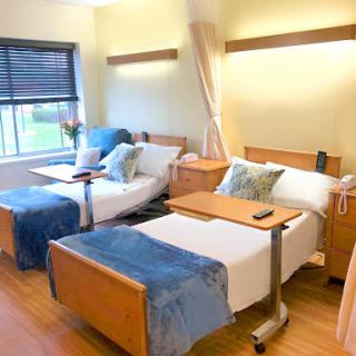 A well-lit hospital room featuring two beds with blue blankets and bedside tables.