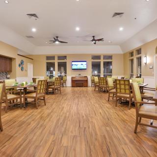 A spacious dining area with wooden flooring, tables, and chairs, featuring ceiling fans and a television on the wall.