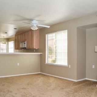 An empty living room with carpet flooring, a large window, and a view into the kitchen area.