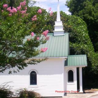 A quaint white church with a green roof and steeple, surrounded by lush trees and pink flowering shrubs.