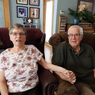 A senior couple sitting in a cozy living room, holding hands and surrounded by family photos.