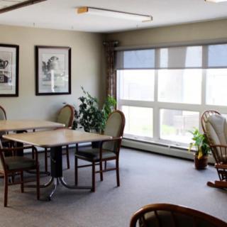 A cozy room with tables and chairs, featuring large windows and plants.