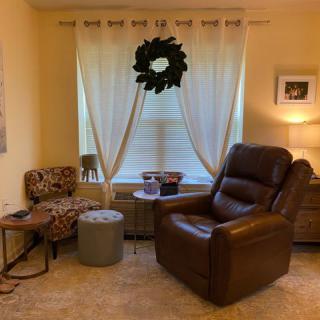 A cozy living room featuring a leather recliner and patterned chair, accented by light curtains and a decorative wreath.