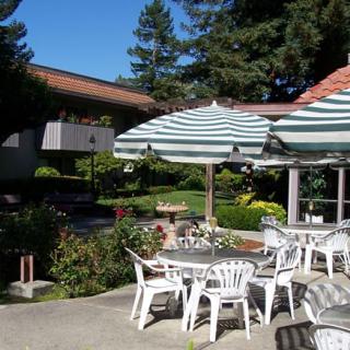 A sunny outdoor patio area with tables, chairs, and striped umbrellas surrounded by a garden.