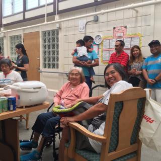 A group of people gathered indoors, including elderly individuals seated and others standing nearby.