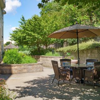 A peaceful patio with outdoor furniture and a large umbrella, surrounded by lush greenery on a sunny day.