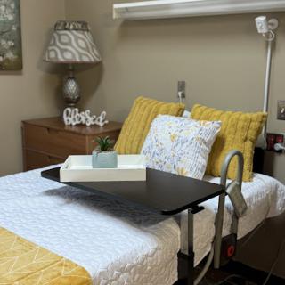 A cozy hospital room featuring a neatly made bed with yellow and white bedding, accompanied by a bedside lamp and a table tray.