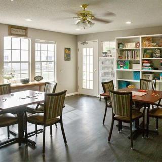 A cozy dining room with wooden tables and chairs, featuring bookshelves and large windows allowing natural light.
