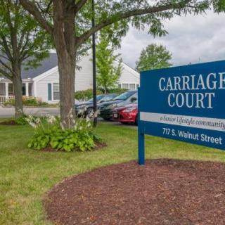 A sign for Carriage Court with a building and parked cars in the background.