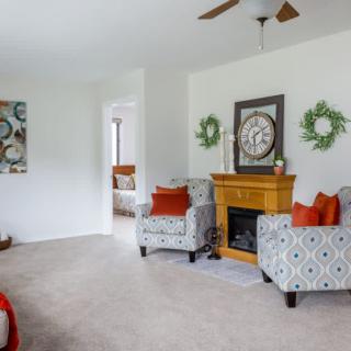 A cozy living room featuring patterned armchairs, a wooden fireplace with a large clock above it, and decorative wreaths on the wall.
