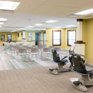 A spacious gym interior featuring exercise equipment, chairs, mirrors, and windows.