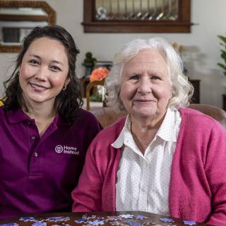 A caregiver and an elderly woman sitting together, smiling in a cozy home setting.