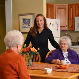 A caregiver interacting with two elderly women sitting at a kitchen table.