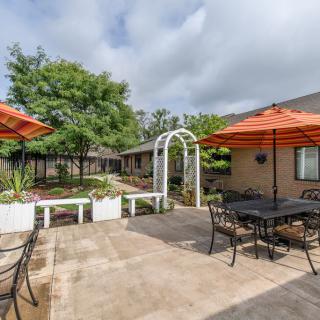 A spacious outdoor patio area with tables, chairs, and orange umbrellas surrounded by greenery and a white archway.