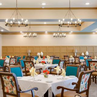 A well-decorated dining room with elegantly set tables and chairs, featuring warm lighting and floral decor.