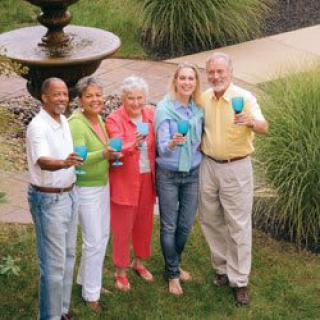 A group of people smiling and holding cups in a garden setting near a fountain.