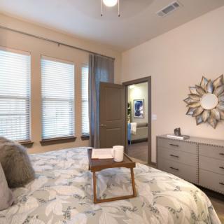 A modern bedroom with a neatly made bed featuring a decorative tray, a dresser with a stylish mirror, and large windows with blinds.