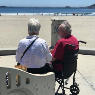 An elderly couple enjoys a peaceful day at the beach, with one seated in a wheelchair.