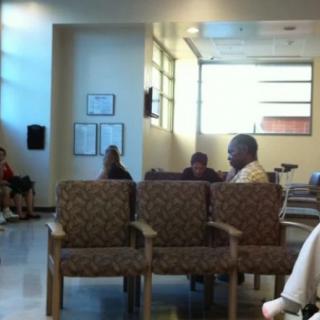 A group of people seated in a waiting room with ample natural light and patterned chairs.