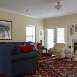 A cozy living room with a blue sofa, armchair, and a coffee table on a patterned rug, featuring a fireplace and artwork on the walls.