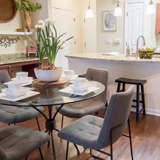 A cozy dining area with a round glass table set for four, featuring a centerpiece plant and modern gray chairs adjacent to a kitchen with bar stools.