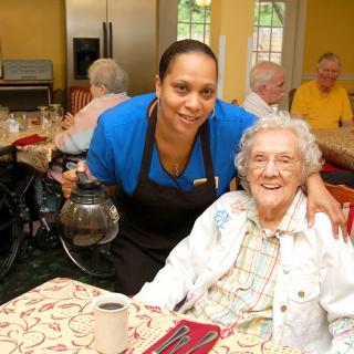 A caregiver serving coffee to a smiling elderly woman in a nursing home setting.