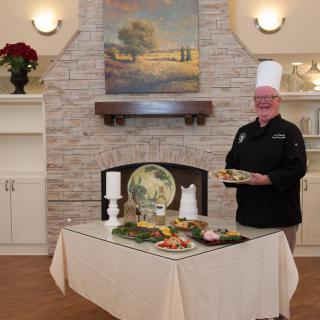 A chef in a black uniform and white hat stands by a table with various dishes, showcasing culinary skills.