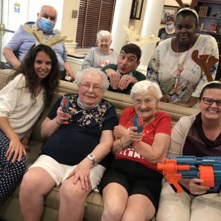 A group of people, including elderly individuals, enjoying a playful moment indoors with toy guns and reindeer antlers.