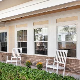 A cozy porch featuring two white rocking chairs beside vibrant plant pots and large windows.