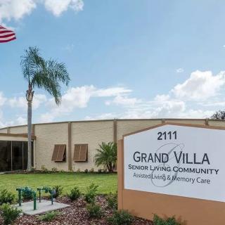 An exterior view of Grand Villa Senior Living Community with a neatly landscaped entrance and an American flag flying nearby.