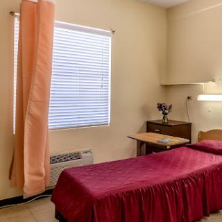 A simple, well-lit room featuring a single bed with a maroon bedspread, a desk, and natural light coming through the window.