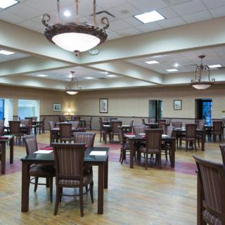 An empty dining hall with wooden tables and chairs, illuminated by chandeliers.