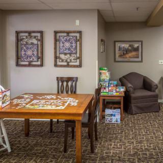 A cozy indoor space featuring a puzzle on a wooden table, surrounded by chairs and an armchair, with artwork adorning the walls.
