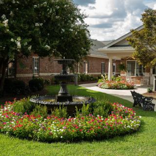 A serene garden with a central fountain, surrounded by colorful flowers and greenery, next to a building.