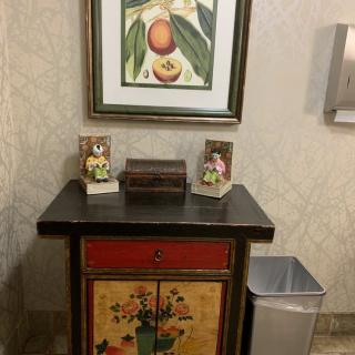 A decorative cabinet with a framed fruit painting above it, flanked by two small figurines.