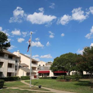 A scenic view of a building complex with a flagpole, surrounded by trees and under a blue sky with fluffy clouds.