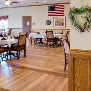 A spacious dining room with neatly arranged tables and chairs, featuring wooden floors and an American flag on the wall.