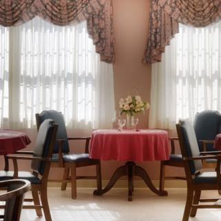 An elegant dining room with round tables, adorned with red tablecloths and flower centerpieces, illuminated by natural light through curtained windows.