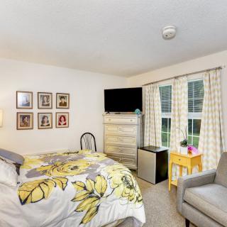 A cozy bedroom with a floral bedspread, a television mounted on a dresser, and natural light coming through the window with plaid curtains.