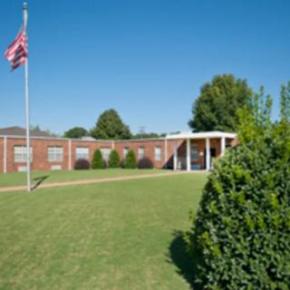 A brick school building with a flagpole and neatly trimmed lawn under a clear blue sky.