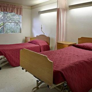 A hospital room with two beds covered in red bedspreads, located near a window with curtains.