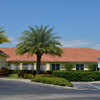 A building with a tile roof, surrounded by palm trees and landscaping, adjacent to a parking lot under a clear blue sky.