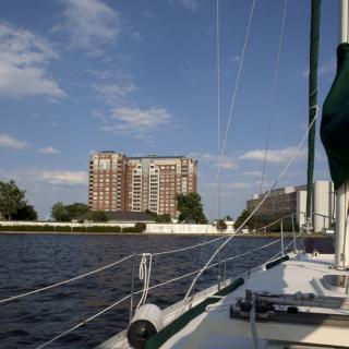 A sailboat cruising on a river with a city building in the background under a partly cloudy sky.